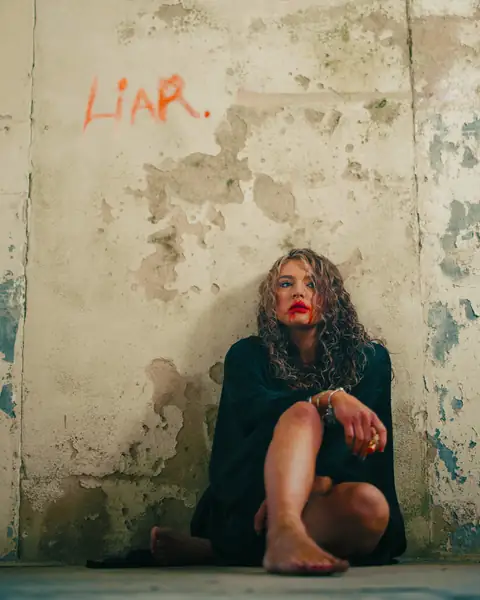 Maddison A sitting against a peeling wall with LIAR written above